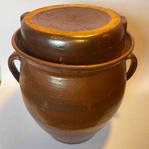 Op de afbeelding: Een bruine keramische pot met een deksel. De pot heeft twee handvatten en een brede opening. Het deksel zit bovenop de pot en heeft een iets verhoogd midden. De pot en het deksel zijn geglazuurd met een glanzende afwerking.