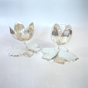 Può includere: Due portacandele color argento a forma di fiori. Ognuno ha una coppa a forma di fiore su uno stelo curvo, appoggiata su una base a forma di foglia. I portacandele sono in metallo lucido e hanno un design dettagliato.