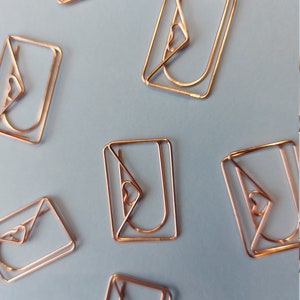 K&ouml;nnte beinhalten: Sechs ros&eacute;goldene Briefumschlaghalter mit Herzdetail. Die Halter sind auf einem hellblauen Hintergrund angeordnet.