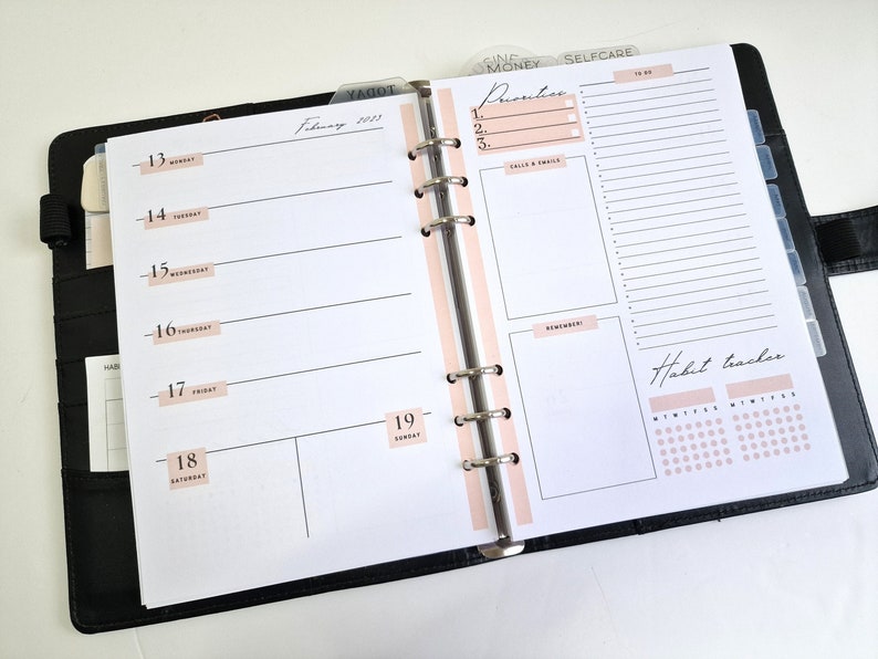 May include: A black planner with a pink tab labelled "February 2023". The planner has a daily layout with space for notes and a habit tracker.