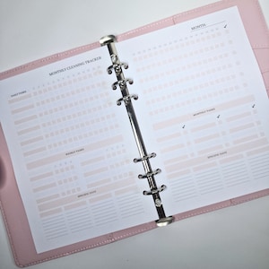 May include: A pink binder with a white page open to a monthly cleaning tracker. The page has sections for daily tasks, weekly tasks, monthly tasks, and specific days. The page is divided into a grid with checkboxes for each day of the month.
