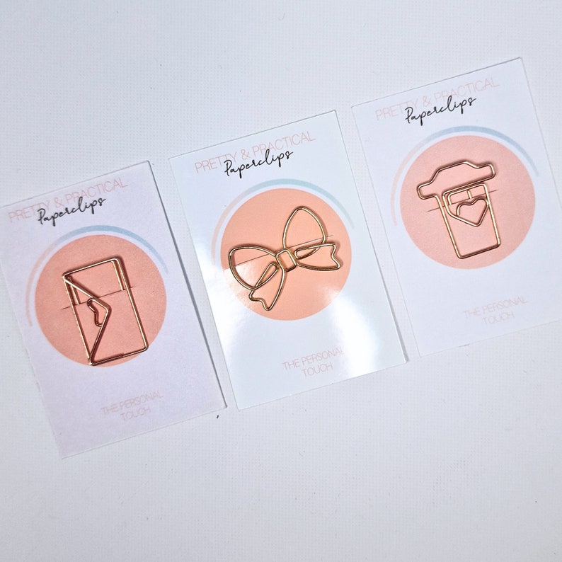 K&ouml;nnte beinhalten: Drei goldene B&uuml;roklammer-Designs auf wei&szlig;en Karten mit rosa Akzenten. Die Designs sind eine Schleife, ein Umschlag und eine Kaffeetasse mit einem Herzen. Die Karten tragen den Text "PRETTY & PRACTICAL Paperclips" und "THE PERSONAL TOUCH".