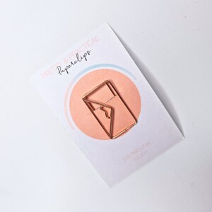 K&ouml;nnte beinhalten: Ein goldener B&uuml;roklammer in Form eines Umschlags mit dem Text "PRETTY & PRACTICAL Paperclips" und "THE PERSONAL TOUCH" auf einer wei&szlig;en Karte mit einem rosa und blauen Kreis.