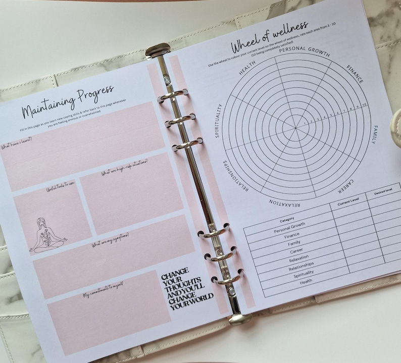 May include: A planner with a pink and white cover and a page with a wheel of wellness chart. The chart has categories for health, personal growth, spirituality, relationships, relaxation, career, finance, and family. The page also has a section for maintaining progress with prompts for what you want to see, what were big, unexpected wins, what are my aspirations, and my commitment to myself. The page also has the text "Change your thoughts and you'll change your world".