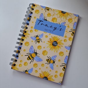 Op de afbeelding: Een geel notitieboek met een spiraalbinding. De omslag heeft een bijen- en zonnebloemmotief met een blauw label waarop "Tracy's Notes" staat.
