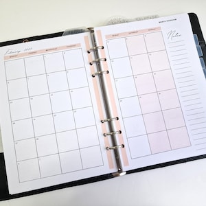 May include: A black planner with a pink and white monthly calendar spread for February 2023. The planner has a ring binding system and includes tabs for different categories, such as "Today", "Selfcare", and "Money".