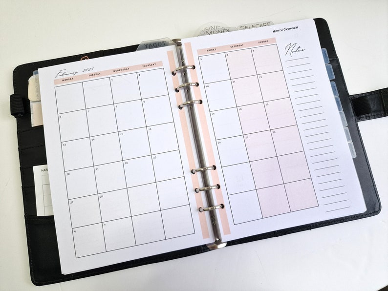 May include: A black planner with a pink and white monthly calendar spread for February 2023. The planner has a ring binding system and includes tabs for different categories, such as "Today", "Selfcare", and "Money".
