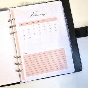 May include: A white planner with a black binder and a pink February calendar page. The calendar page has the days of the week labeled and the numbers 1 through 28.