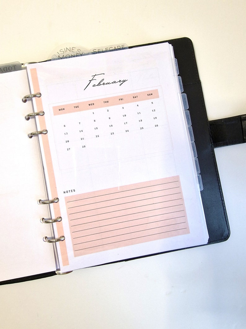 May include: A white planner with a black binder and a pink February calendar page. The calendar page has the days of the week labeled and the numbers 1 through 28.