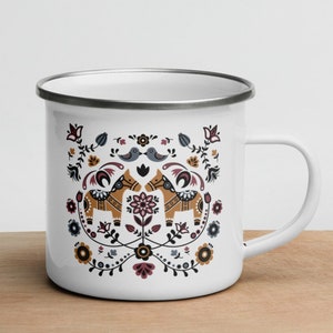 May include: White enamel mug with a silver rim featuring a Scandinavian folk art design. The design includes two stylized horses, birds, and floral patterns in shades of red, blue, yellow, and black.