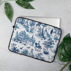 Op de afbeelding: Een laptoptas met een blauw en wit toile de Jouy-patroon. Het ontwerp toont scènes van mensen, gebouwen en bomen. De tas heeft een zwarte rand en ligt op een witte ondergrond met groene bladeren.