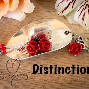 May include: A silver spoon-shaped keychain with red rose accents and a lobster clasp. The spoon features a small hat design with red roses and a leaf. The word "Distinction" is written on the wooden surface.