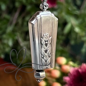 May include: A silver-toned pendant with a floral design. The pendant has a rectangular shape with vertical lines and a decorative floral motif. A small, round bead hangs from the bottom. The background is blurred with green foliage and pink flowers.