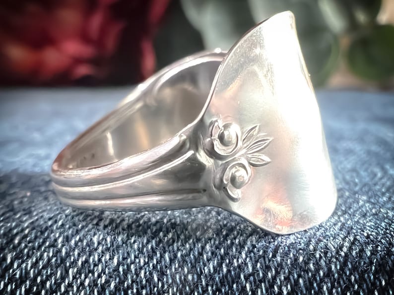 May include: A silver ring made from a repurposed spoon with a floral design. The ring is on a blue denim background.