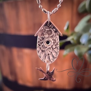 May include: A silver birdhouse pendant necklace. The pendant is crafted from a spoon, featuring floral details and a small bird charm. The necklace hangs on a ball chain, with a blurred background of greenery and a wooden barrel.