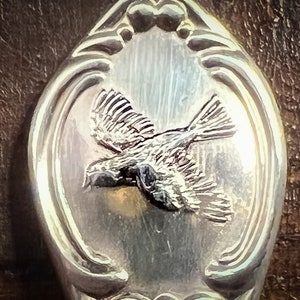 May include: A silver spoon with an ornate design featuring a bird in flight. The bird is detailed with feathers and has a long tail.