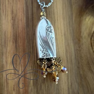 May include: Silver pendant necklace featuring a repurposed spoon bowl with floral detailing. The pendant is adorned with three gold-colored bee charms and purple crystal beads. The necklace hangs on a silver chain.