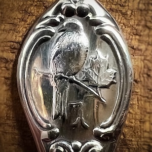 May include: A silver spoon with an ornate design featuring a bird perched on a branch with leaves. The spoon is engraved with the letter "K".