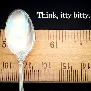 May include: A silver teaspoon with a floral design on the handle is measured against a wooden ruler. The ruler shows the spoon is just over 5 inches long. The text "Think, itty bitty." is written above the ruler.