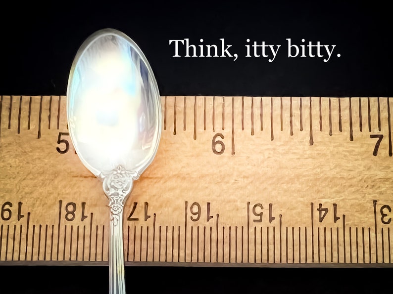 May include: A silver teaspoon with a floral design on the handle is measured against a wooden ruler. The ruler shows the spoon is just over 5 inches long. The text "Think, itty bitty." is written above the ruler.