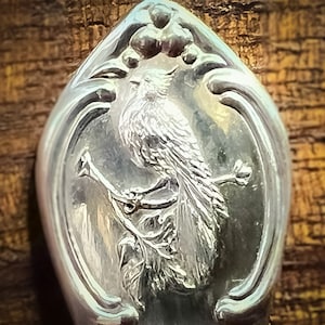 May include: A silver spoon with an ornate design featuring a bird perched on a branch. The bird has detailed feathers and is surrounded by floral motifs.