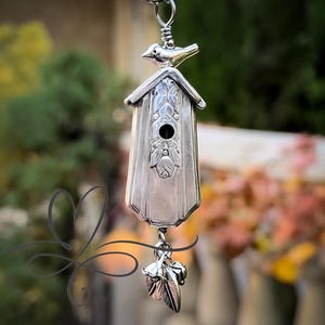 Puede incluir: Colgante plateado con forma de casita para pájaros y un pequeño pájaro en la parte superior. La casita tiene detalles florales y un dije de hoja. Cuelga de una cadena plateada. El fondo borroso muestra vegetación.