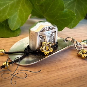 May include: A silver-toned keychain featuring a miniature building charm with geometric details, resting on a spoon-shaped base. A gold-colored flower accentuates the design, with a matching flower charm on the chain. The keychain is set against a wooden surface.