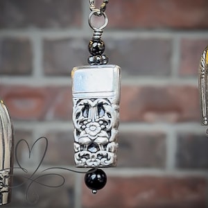 Puede incluir: Primer plano de tres colgantes plateados con intrincados diseños florales y geométricos. Los colgantes cuelgan sobre un fondo de ladrillo borroso. Cada colgante tiene una forma única, incluyendo una campana y un diseño rectangular con una cuenta negra.