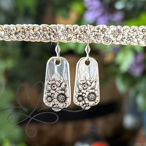 May include: Silver-toned earrings with a floral design. The earrings are rectangular with rounded corners and feature a detailed pattern of flowers. The earrings are suspended from a decorative silver bar with a floral motif.