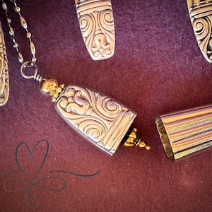 May include: A silver pendant necklace with a bell-shaped charm, featuring ornate scrollwork and gold accents. The necklace is displayed on a deep red-purple surface, with additional silverware pieces in the background.