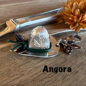 May include: A silver-toned metal keychain shaped like a miniature hat, featuring a dark green leather cord band, a small embossed flower design, and additional dangling charms including a copper-colored flower and leaf, with the word Angora below.