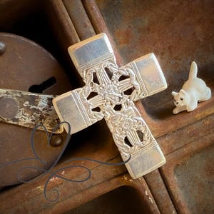 Puede incluir: Una cruz de plata con un diseño intrincado, descansando sobre una superficie metálica oxidada. Una pequeña figurita de gato de cerámica blanca está en el fondo.