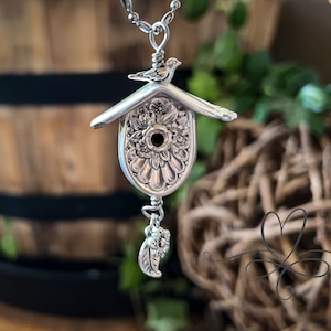 May include: A silver birdhouse pendant necklace. The pendant is crafted from ornate silverware, featuring a bird perched on the roof and a floral design. A small charm hangs below. The necklace is suspended from a delicate chain.