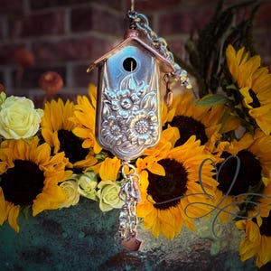 May include: A silver birdhouse charm with a floral design hangs from a chain. The charm is surrounded by yellow sunflowers and white roses.