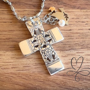 Puede incluir: Un collar con colgante de cruz plateada con intrincados detalles florales. La cruz cuelga de una cadena de cuentas, con un pequeño dije y una perla. El collar se muestra sobre una superficie de madera.