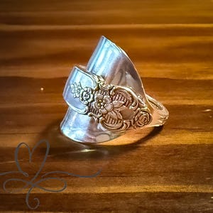 May include: A silver-toned ring crafted from a repurposed spoon, featuring a prominent floral embossed pattern on the handle section that wraps around the finger. The metal has a polished, reflective finish with intricate detailing.