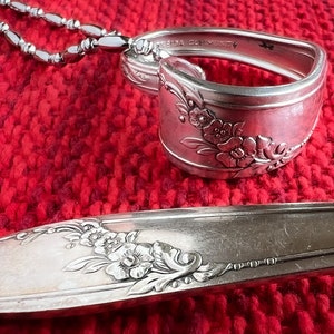 May include: A silver spoon necklace with a floral design. The spoon is bent into a heart shape and has a chain attached to it. The spoon is lying on a red knitted surface.