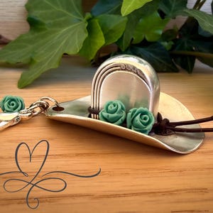 May include: A silver-toned cowboy hat keychain with three turquoise rose accents. The hat has a curved brim and a decorative top. A silver clasp and leather straps with beads complete the design. The keychain is on a wooden surface.