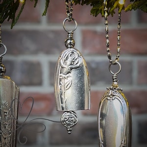 May include: Silver bell ornaments with floral designs hang from a green branch against a brick background. The bells are made from repurposed silverware, with intricate rose and leaf details. Decorative and unique.