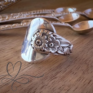 May include: A silver ring crafted from a repurposed spoon, featuring a polished bowl section and a handle segment embossed with a detailed floral pattern. The ring is positioned in front of several matching silver spoons with ornate handles.