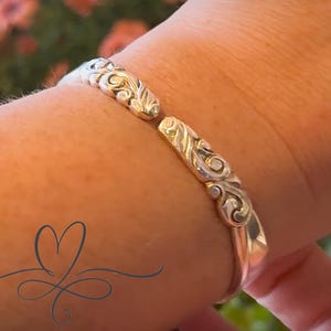 May include: A silver-colored cuff bracelet with an ornate, swirling design. The bracelet is open, revealing the wrist. The intricate pattern features floral and scroll-like elements. The background is blurred, with hints of green and pink.