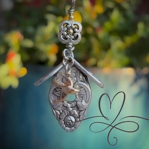 May include: A handcrafted silver-toned birdhouse pendant with a gold-colored bird charm. The pendant is suspended from a decorative floral-shaped bail. The birdhouse is made from a repurposed spoon, and the background is blurred.