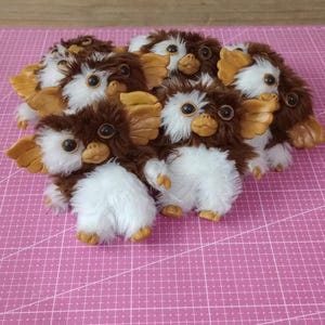 May include: A group of plush Gremlins toys. Each toy features brown fur, white bellies, large ears, and big eyes. The toys are arranged on a pink cutting mat.