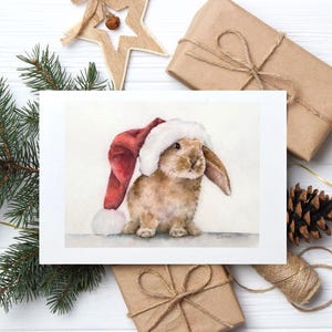 May include: A watercolor painting of a brown bunny wearing a red and white Santa hat. The bunny is the focal point, set against a white background. The image is surrounded by Christmas decorations, including wrapped gifts, pine needles, and a star ornament.