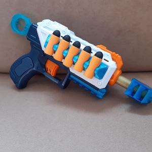 Puede incluir: Una pistola bláster de juguete en tonos azules, blancos y naranjas. El juguete tiene un mango, un cañón y una serie de dardos de espuma naranja con puntas negras. El bláster está diseñado para el juego de los niños y es de plástico.