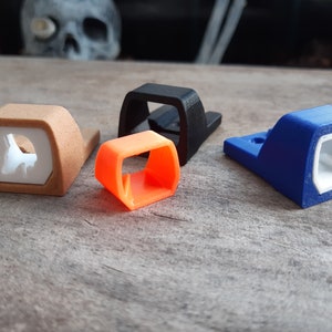 Puede incluir: Cuatro objetos impresos en 3D en varios colores: beige, naranja, negro y azul. Cada objeto tiene una forma única con un interior hueco. Los objetos beige y azul tienen un interior blanco. Están dispuestos sobre una superficie de madera envejecida.