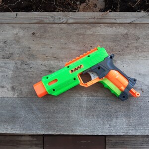 May include: A green and orange toy gun with the word "Radd" printed on the side. The toy gun is made of plastic and has a black handle. The toy gun is lying on a wooden surface.