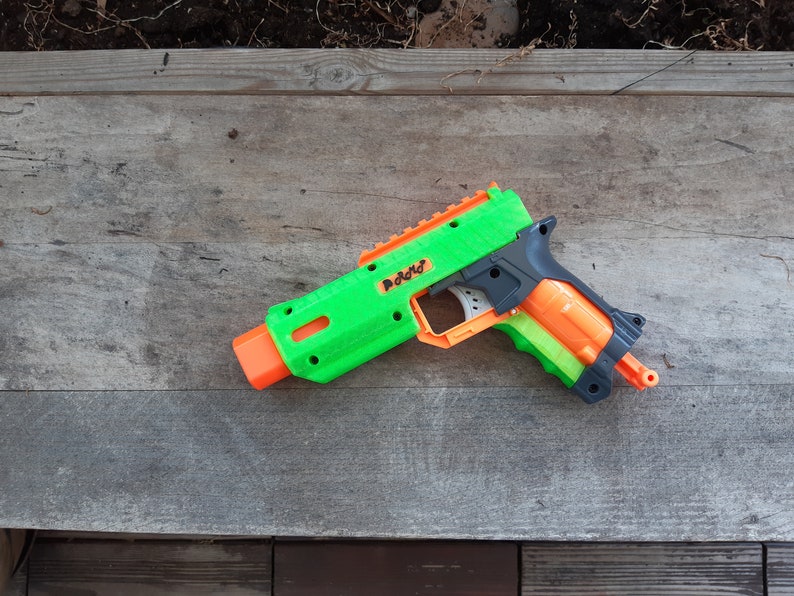 May include: A green and orange toy gun with the word "Radd" printed on the side. The toy gun is made of plastic and has a black handle. The toy gun is lying on a wooden surface.