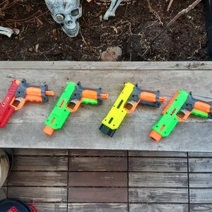 May include: Four toy blasters in red, green, and yellow with orange accents are arranged on a wooden surface. The blasters have a pistol-like design, with a trigger and barrel. A decorative skull is visible in the background.
