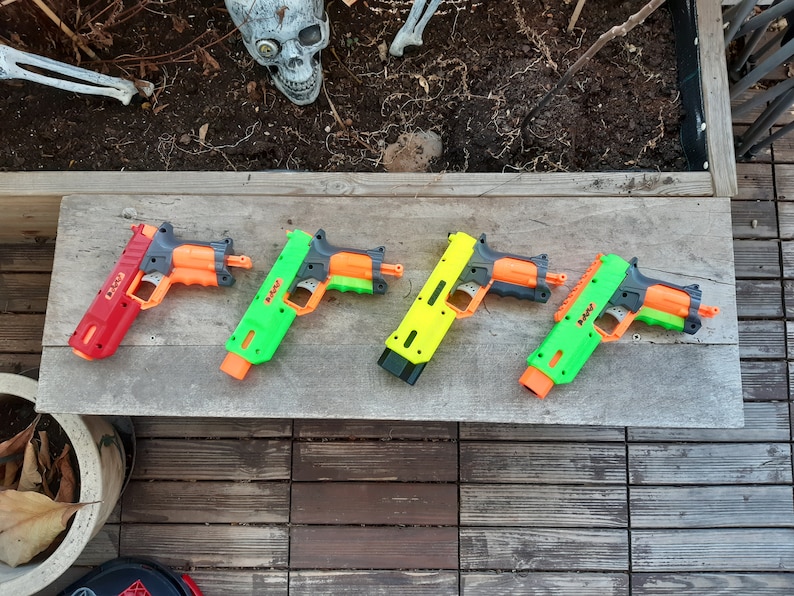 May include: Four toy blasters in red, green, and yellow with orange accents are arranged on a wooden surface. The blasters have a pistol-like design, with a trigger and barrel. A decorative skull is visible in the background.